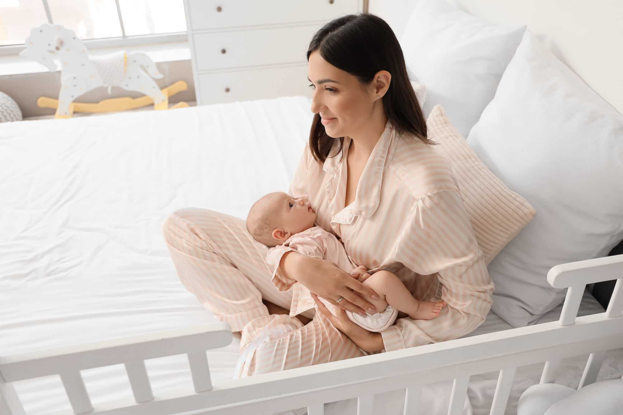 Happy mother with cute little baby sitting on bed in room