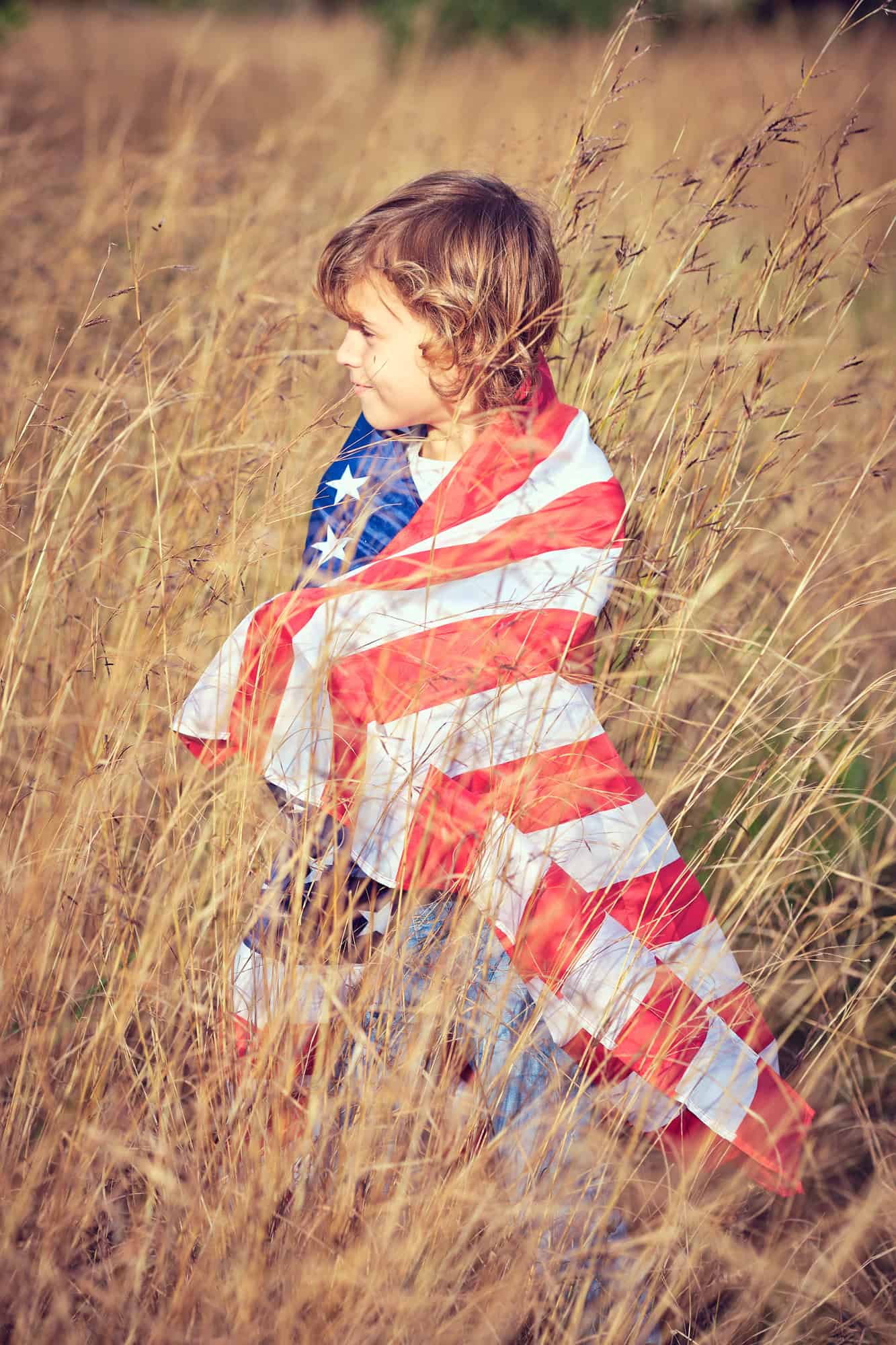 Side view of cute preteen boy wrapped in national flag of United States of America standing in rye meadow