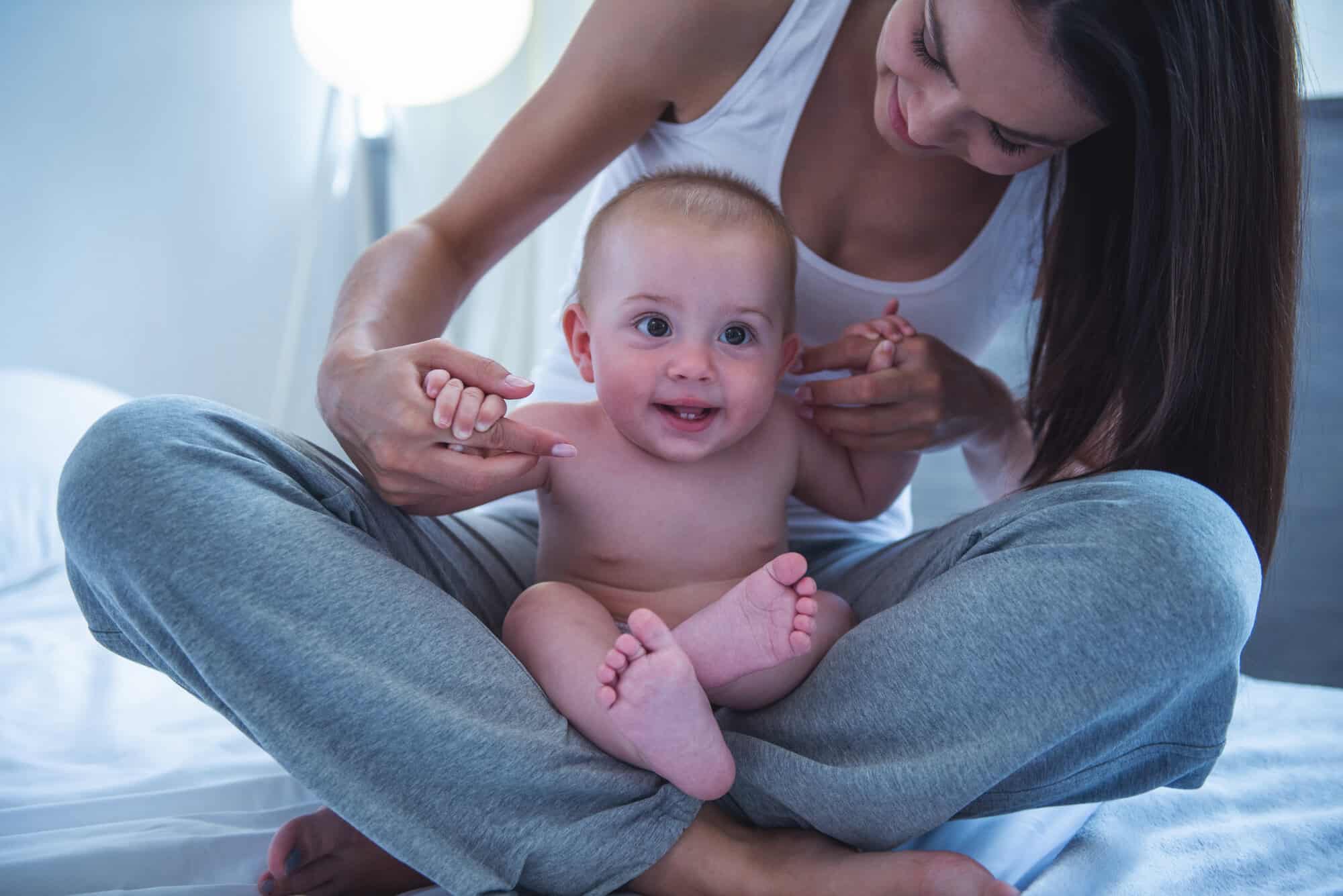 Portrait of charming naked little baby looking away and smiling while sitting on his mother's knees at home. Mom is looking at her child