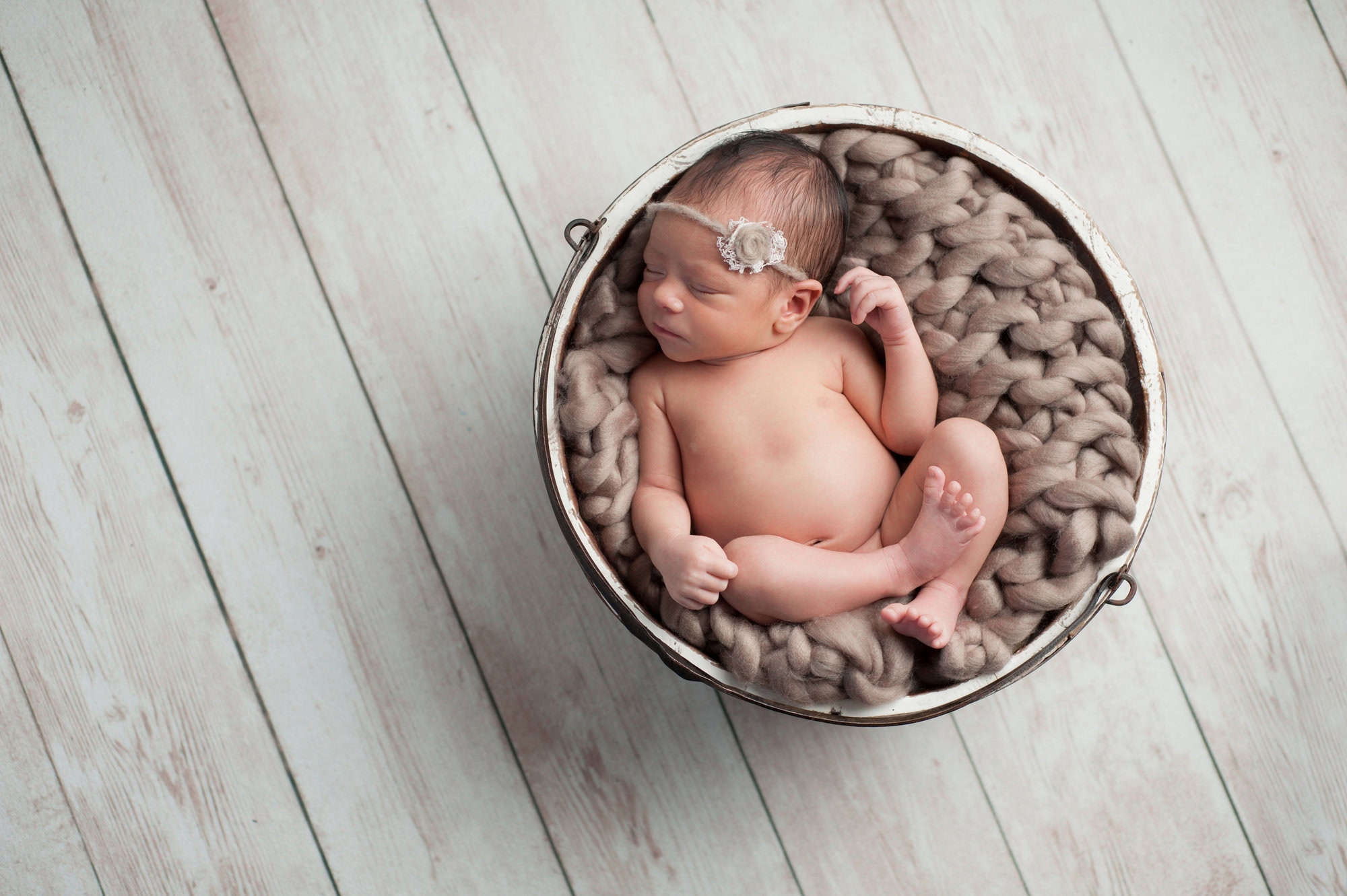 Two week old newborn baby girl sleeping in a wooden bucket. She is wearing a taupe colored headband. Shot in the studio on a white wood background.