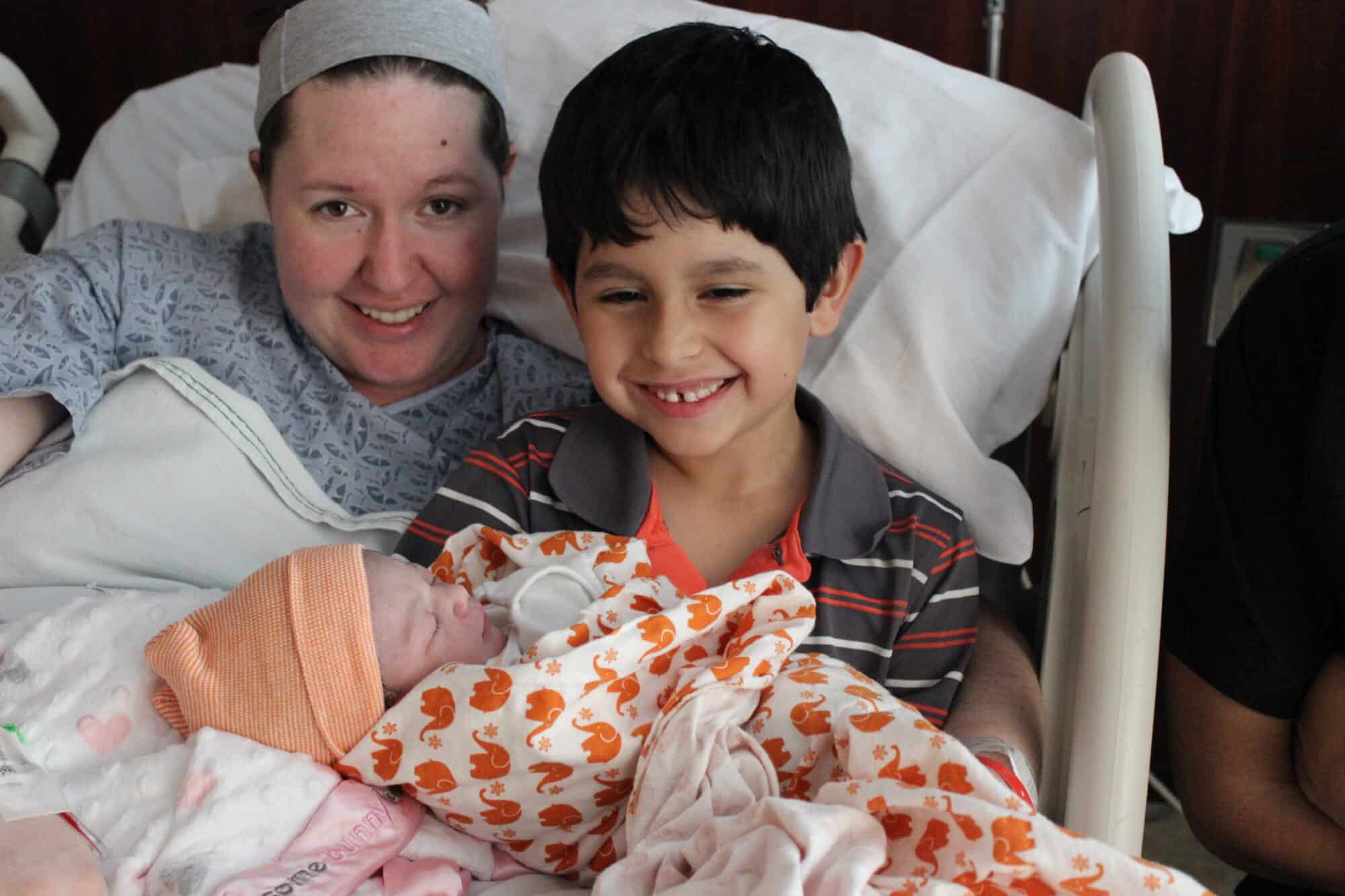 Mom and son in hospital bed with swaddled newborn baby being held by older brother.