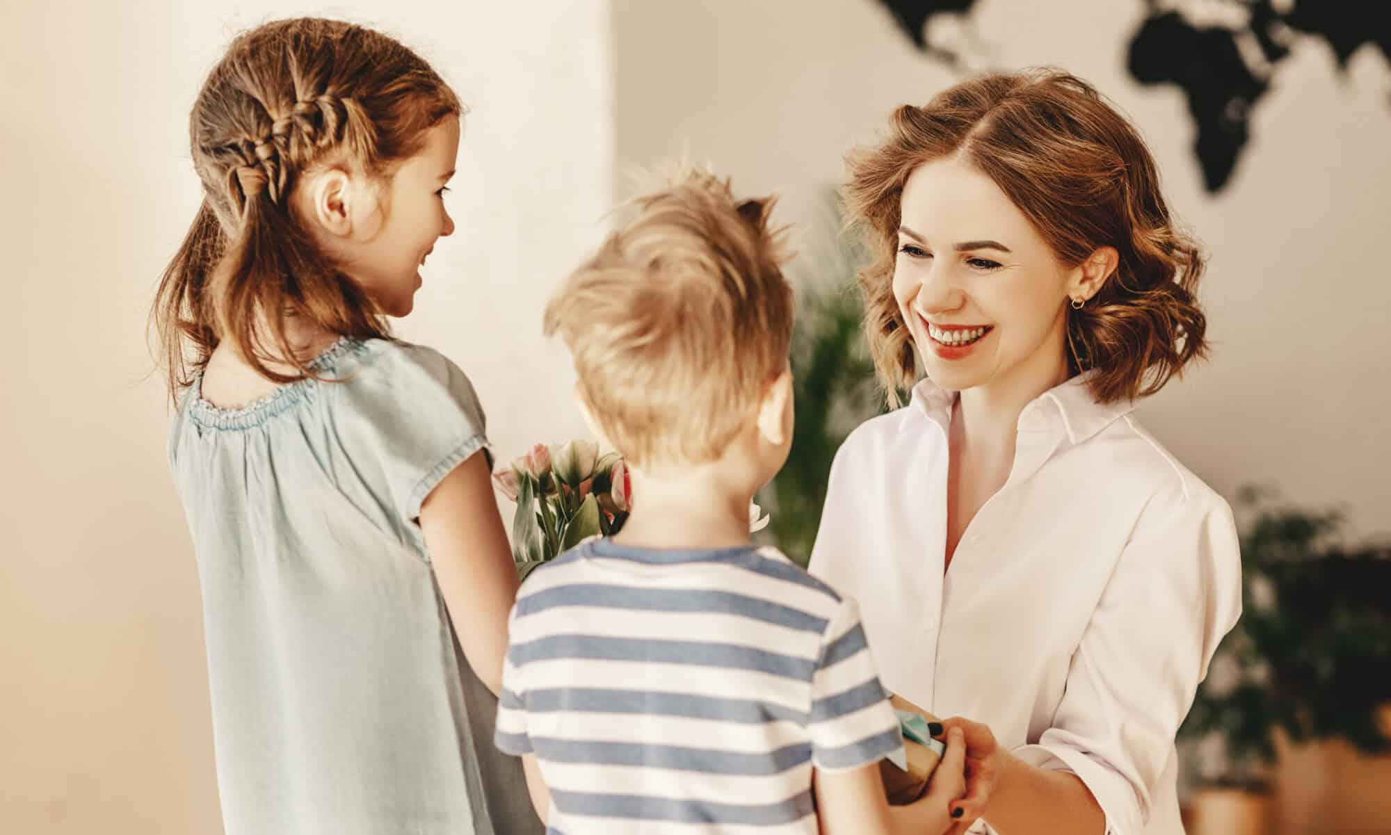 Happy Mothers Day! Children boy and girl congratulate smiling mother and give her flowers bouquet of tulips during holiday celebration in kitchen at home