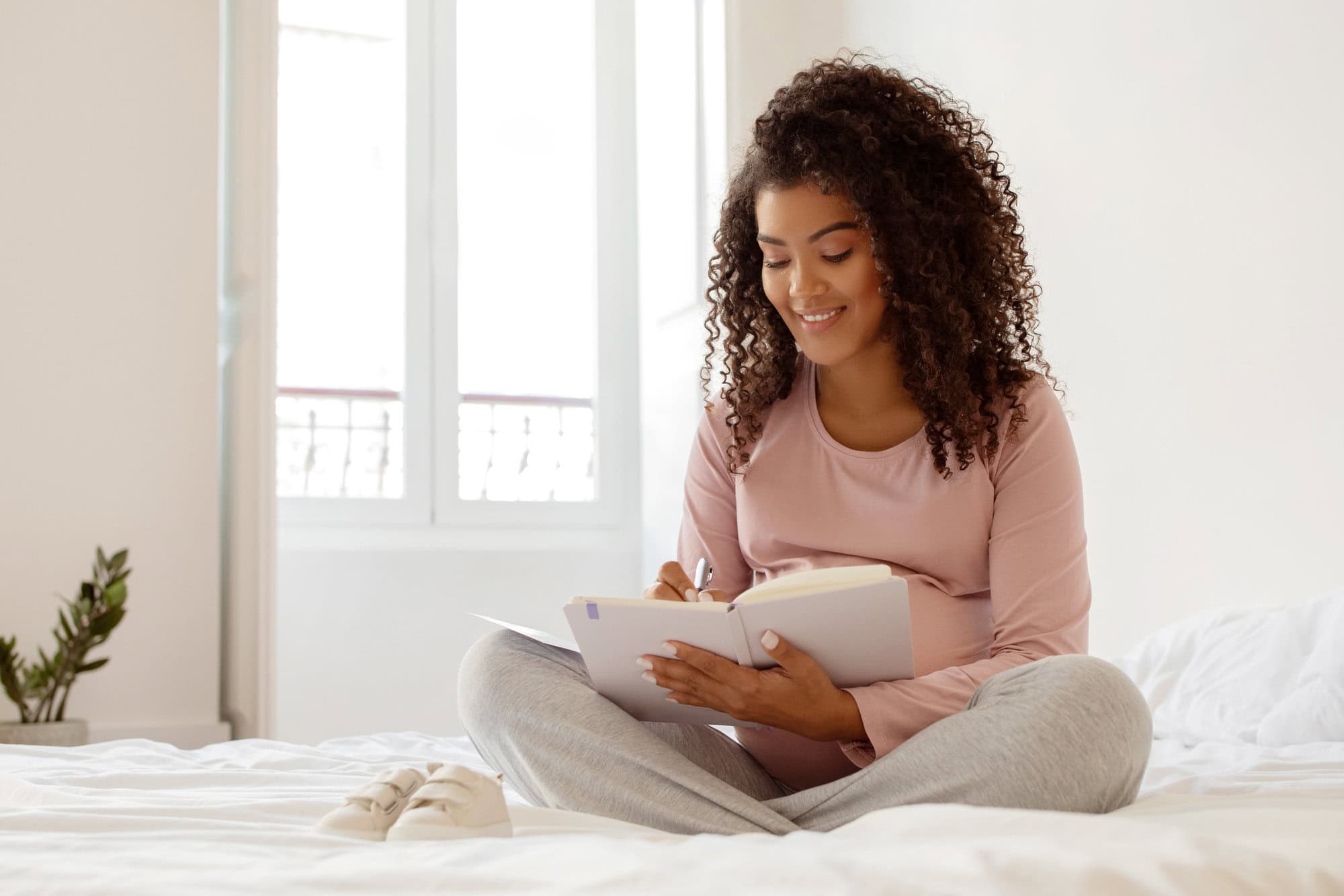 Beautiful black pregnant woman writing in notepad while sitting on bed at home, smiling expectant mother capturing precious pre-birth moments, surrounded by peaceful bedroom ambiance