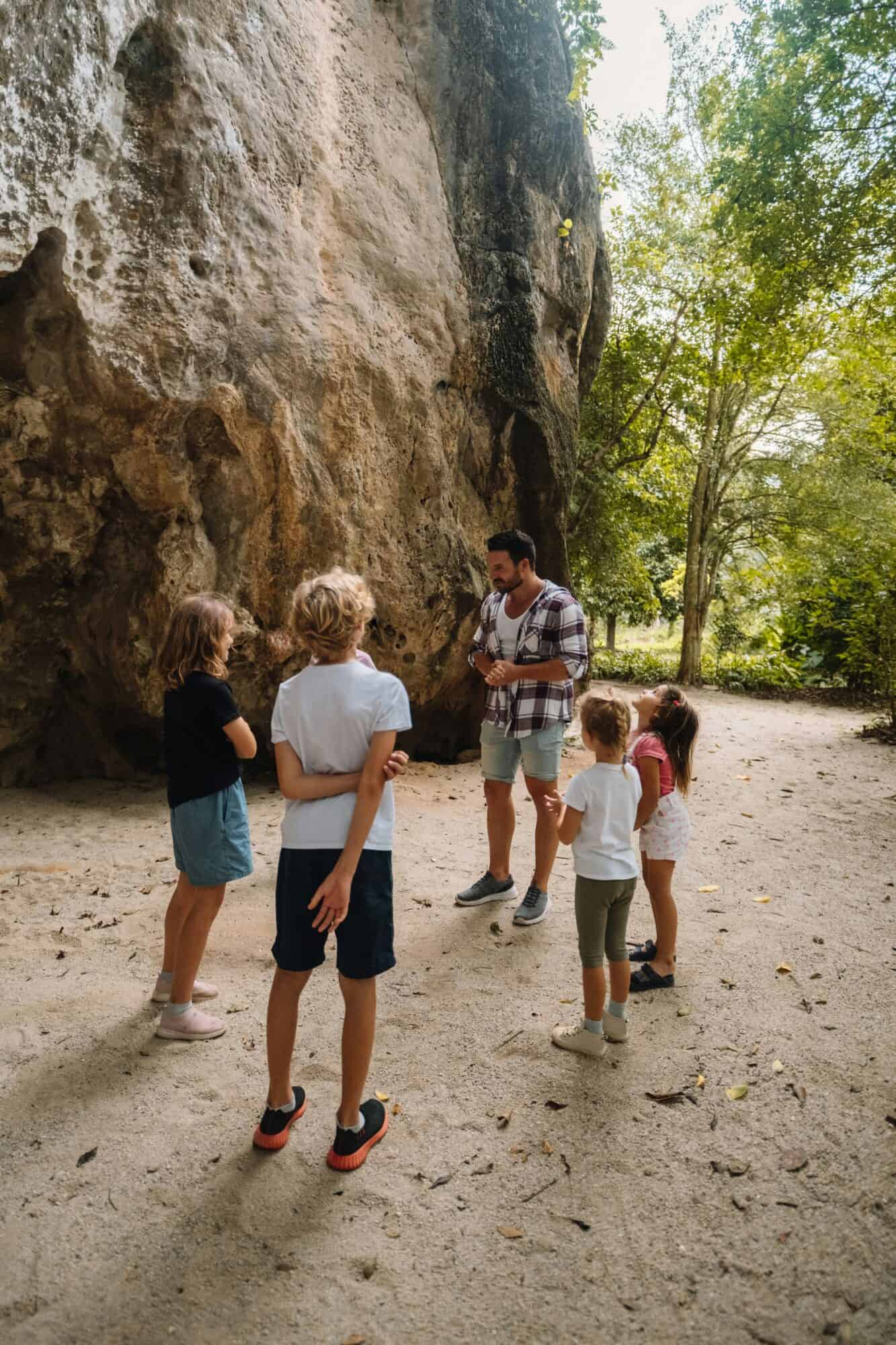 Dad standing with his kids outdoors talking with them. They are dressed for summer on a forested trail talking in a group.