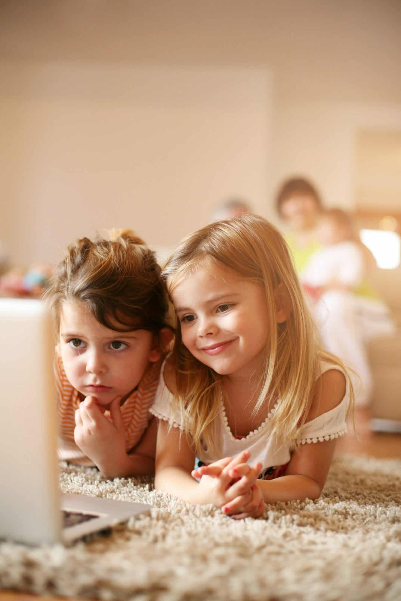 Two sisters laying on the living room floor looking at a laptop.