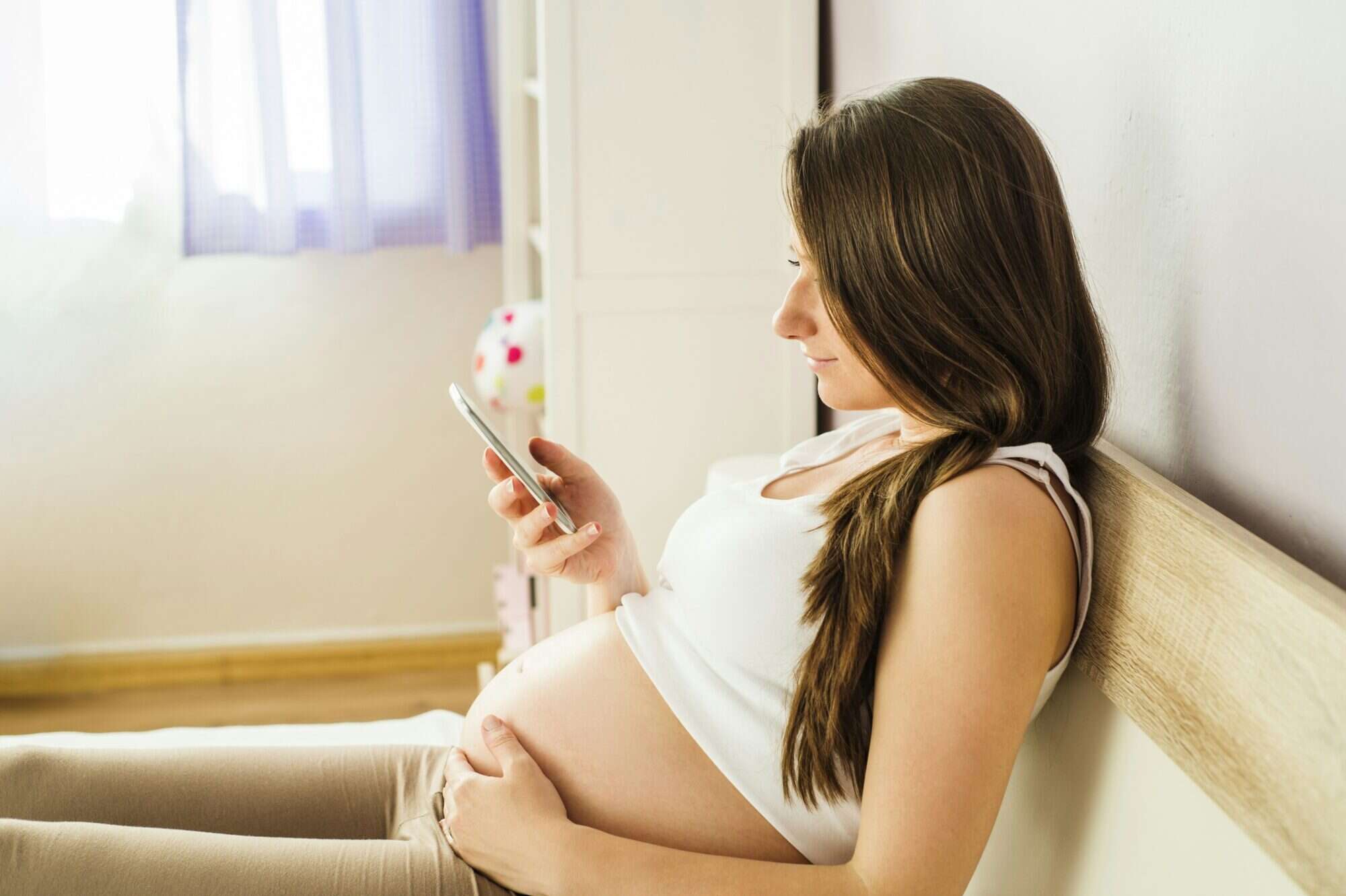 Pregnant mom sits on bed looking at smart phone.