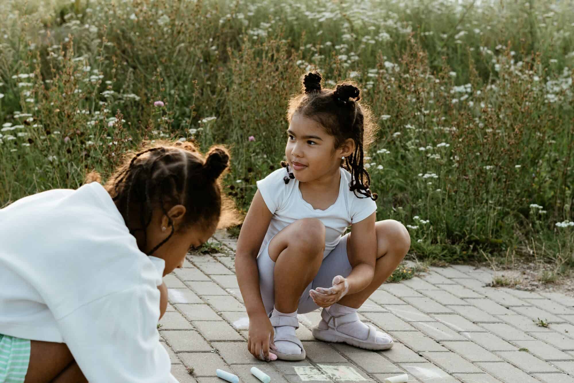 Two girls color with chalk on a sidewalk outdoors.