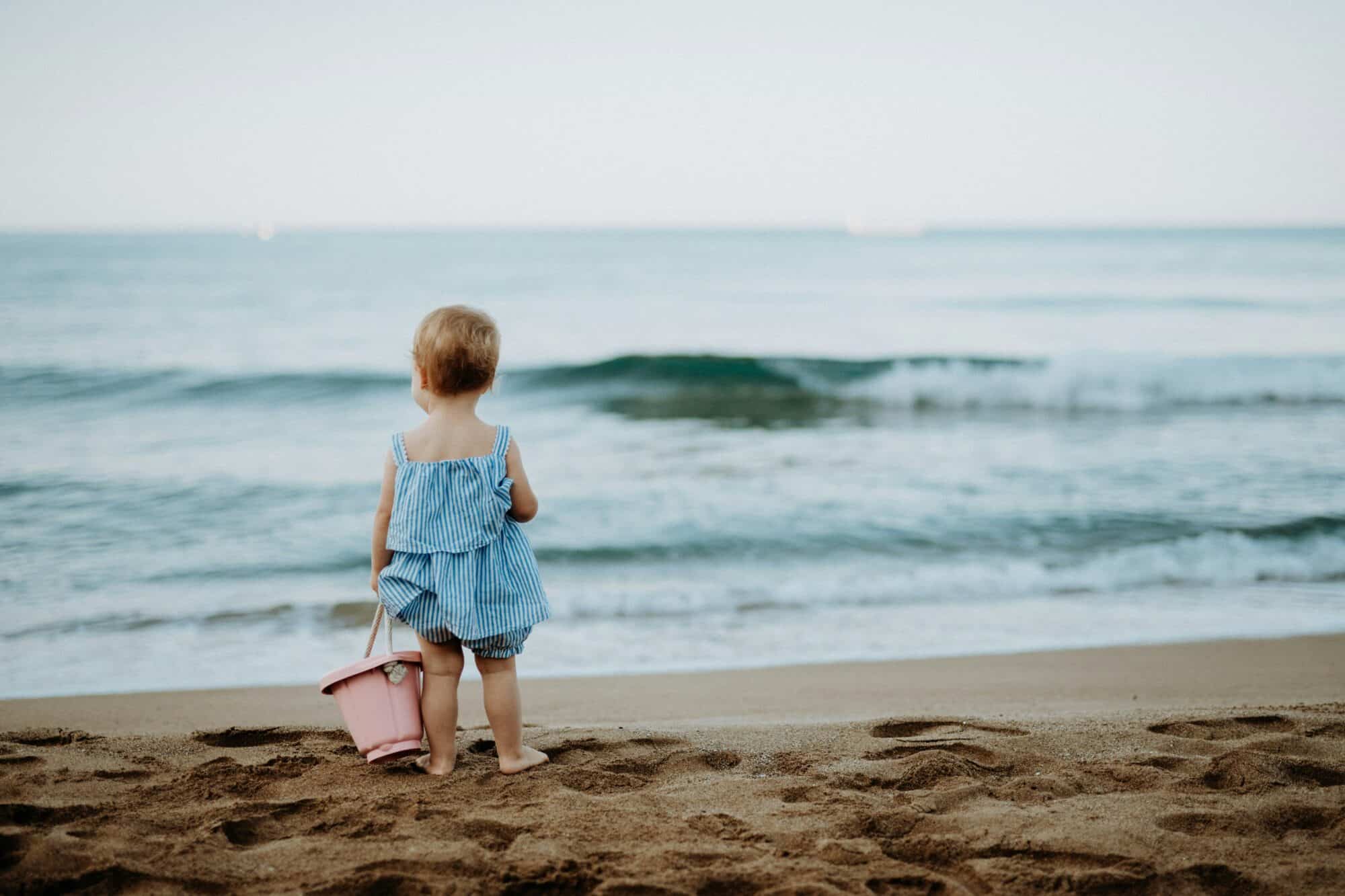 toddler on the beach with pink pail and blue and white striped romper