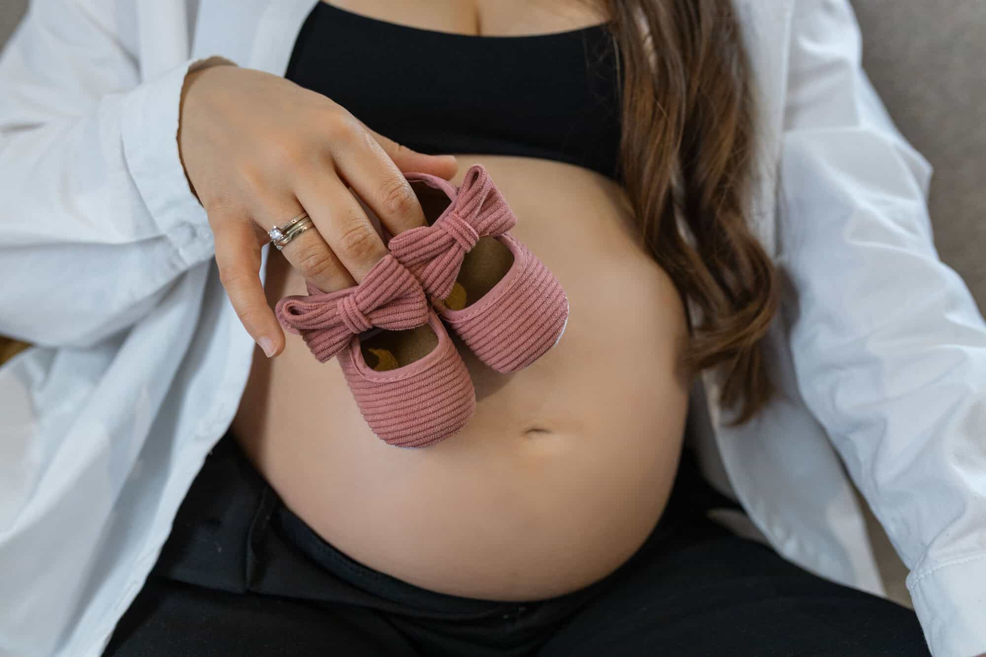 Pregnant woman sitting on sofa with baby shoes resting on baby bump. High quality photo